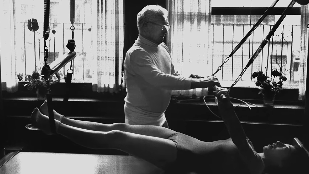 Joseph Pilates assists a woman with an exercise on a spring-based Pilates apparatus.