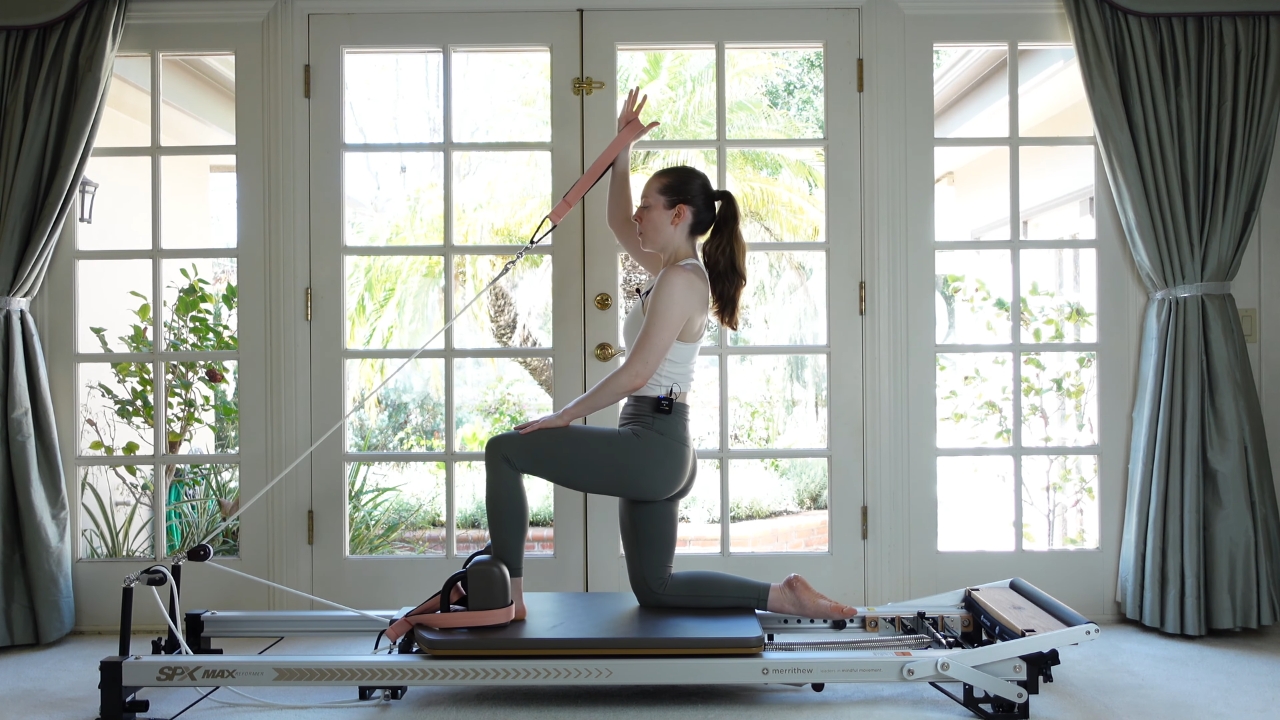 Woman kneels on a Reformer Pilates machine with one arm raised against a resistance strap.