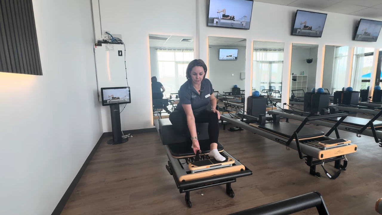 A woman adjusts a Pilates reformer in a studio equipped with exercise screens.