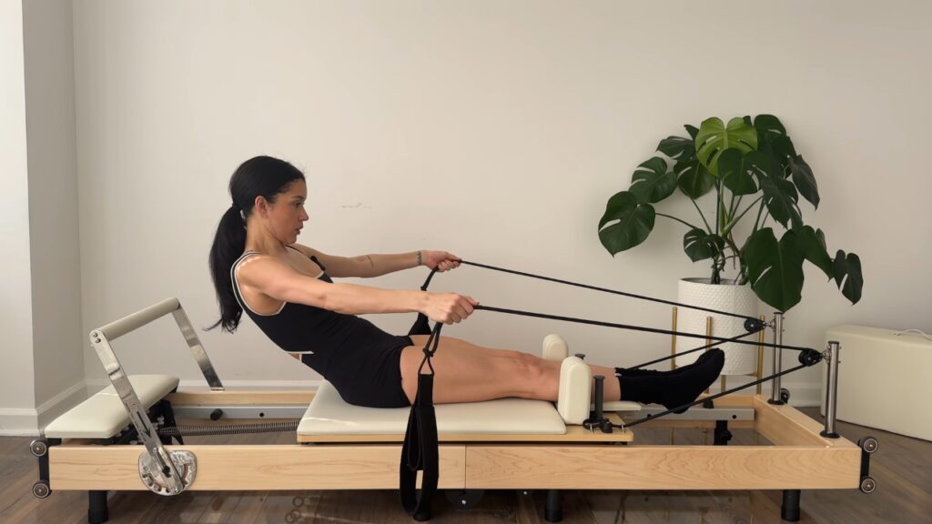 Woman performs Reformer Pilates with resistance straps on a wooden machine.